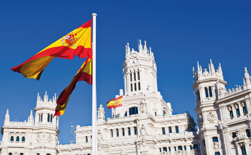 Spanish flag waving in front of majestic Neo-Classical palace under clear blue sky.