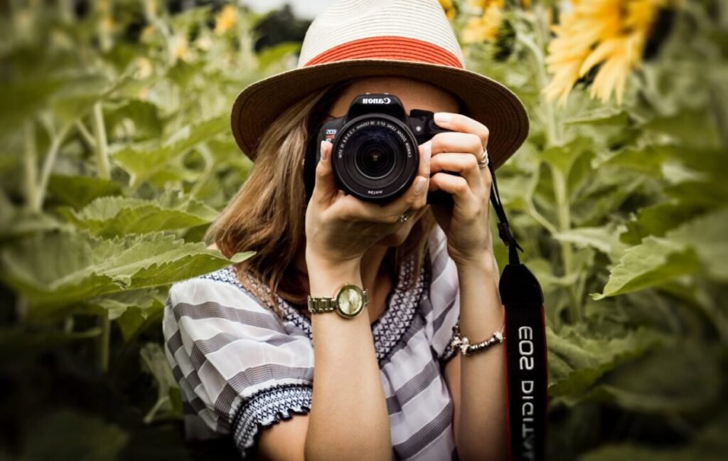 Woman wearing a straw hat taking a photo with a Canon camera, surrounded by sunflowers.