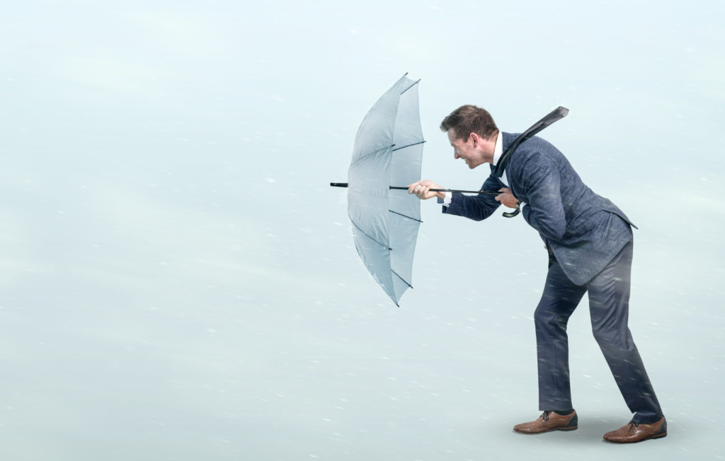 A man in a suit leaning forward and struggling against strong wind while holding an inverted umbrella.