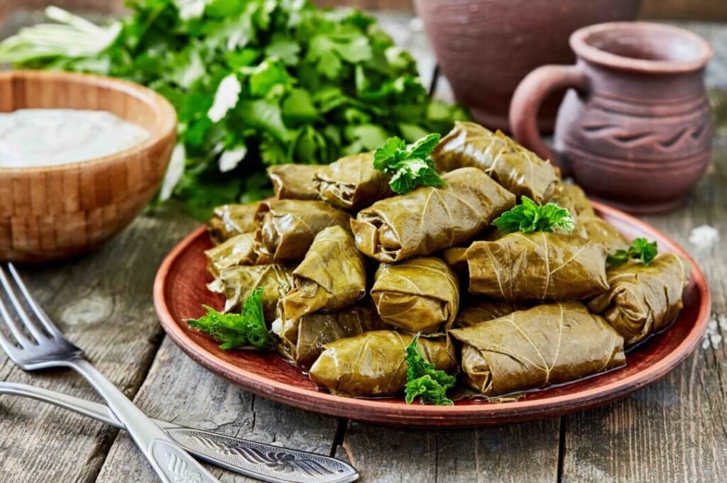 Stuffed grape leaves on a terracotta plate garnished with parsley, accompanied by a bowl of sauce and a bunch of fresh herbs on a rustic wooden table.