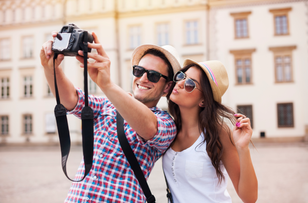 A smiling young couple in summer attire taking a selfie with a DSLR camera in front of a historic building.