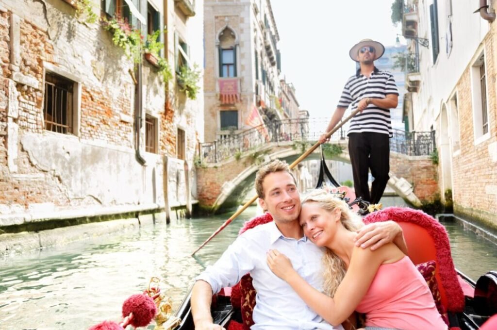 A smiling couple embraces on a gondola ride with a gondolier steering in the background, in a sunny Venetian canal with historic buildings and a small bridge.