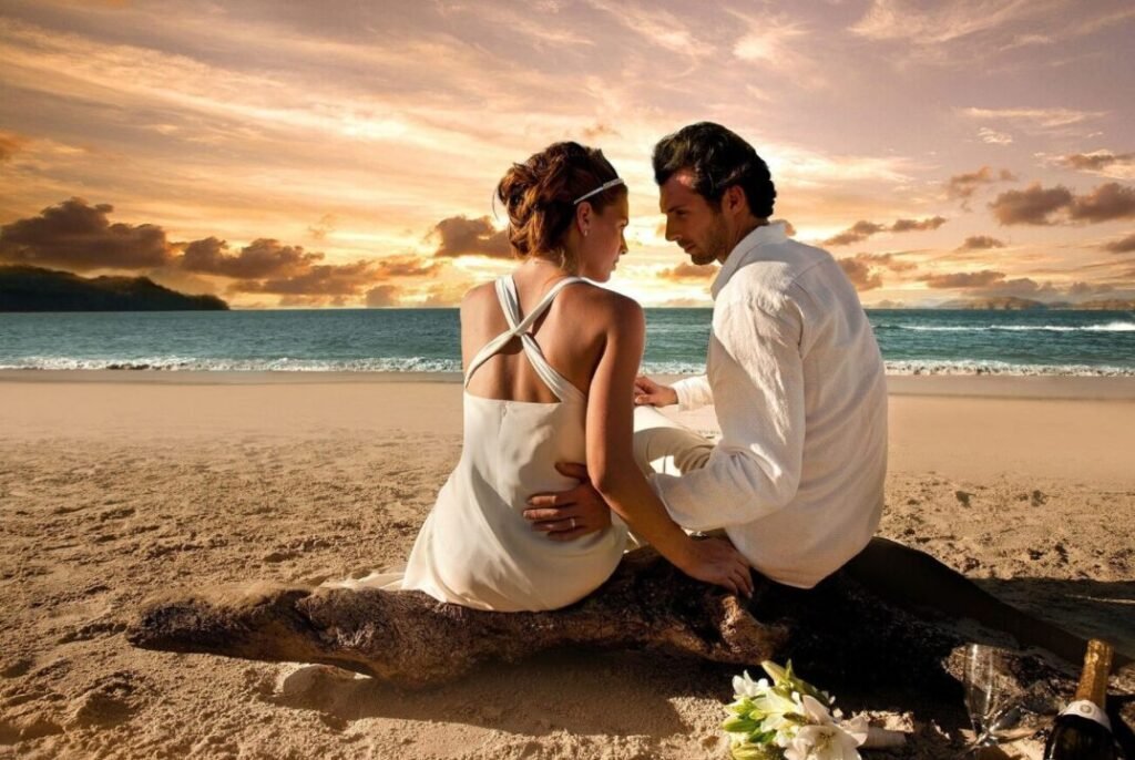 A couple sits closely together on a driftwood log on the beach at sunset, with a bouquet of white flowers and a bottle of champagne beside them.