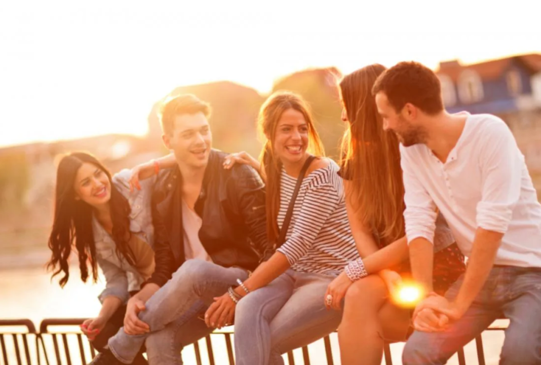 Friends sharing laughs during golden hour chat on urban railing, sunset backdrop.