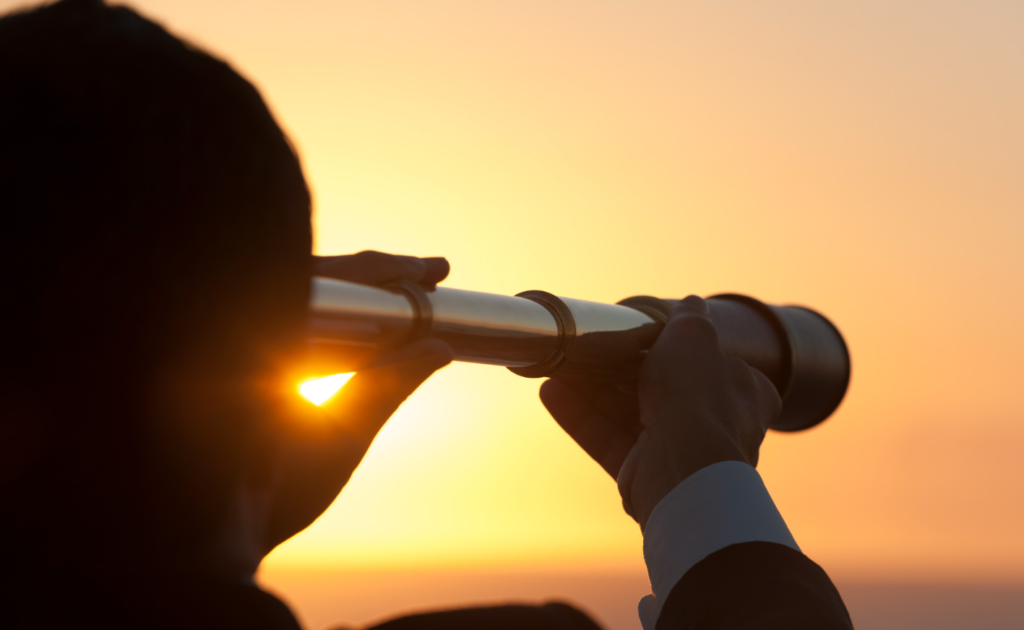 A person in silhouette looking through a telescope during sunset, with the sun casting a warm glow in the background.