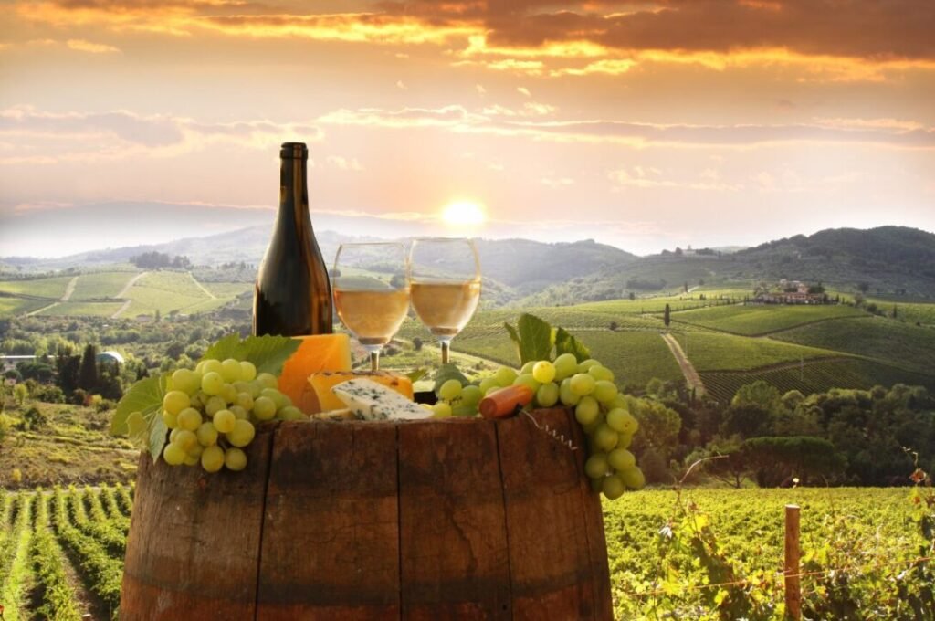 Alt text: A picturesque sunset view over a vineyard with a bottle and two glasses of white wine, grapes, cheese, and bread on a wooden barrel in the foreground.