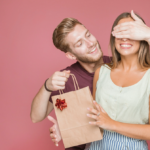 Happy couple exchanging surprise gift, man covers womans eyes with gift bag.