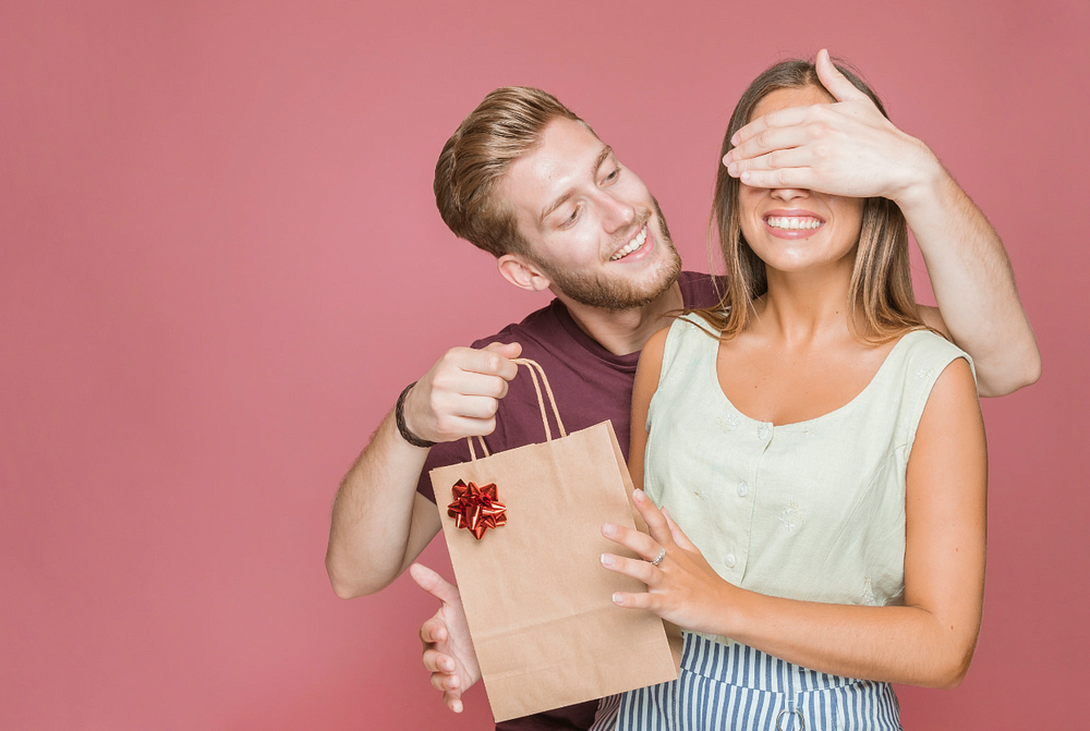 Happy couple exchanging surprise gift, man covers womans eyes with gift bag.