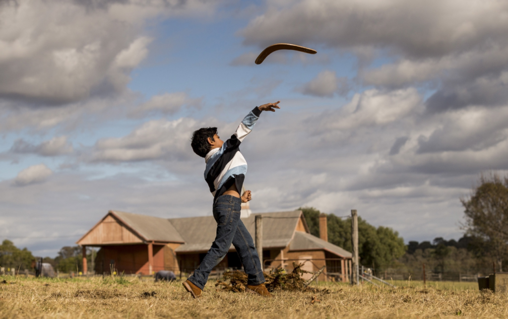 A boy throwing a boomerang in an open field with farm buildings in the background and a cloudy sky above.