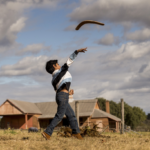 Person throwing boomerang in rural field under cloudy sky.