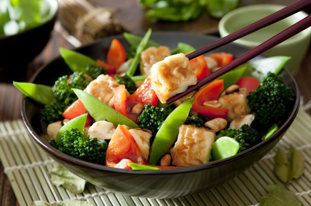 A bowl of stir-fried tofu with broccoli, snap peas, and red bell peppers, picked up with dark chopsticks, on a bamboo mat.