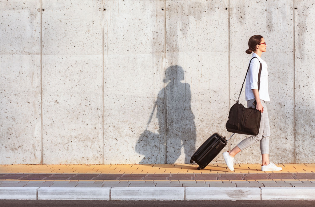 City traveler with rolling luggage walking along urban sidewalk, casting long shadow on wall.