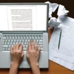 Focused writing process with laptop, documents, and green pen on wooden desk.
