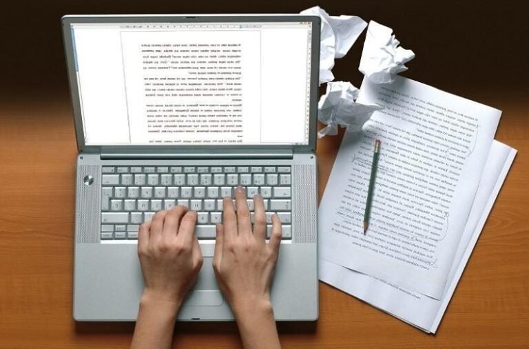 Focused writing process with laptop, documents, and green pen on wooden desk.