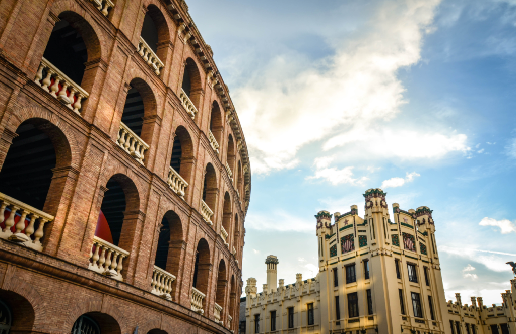 A view of two contrasting buildings against a partly cloudy sky; on the left is a classic brick building with arched openings, and on the right is an ornate, cream-colored building with decorative elements and sculptures.