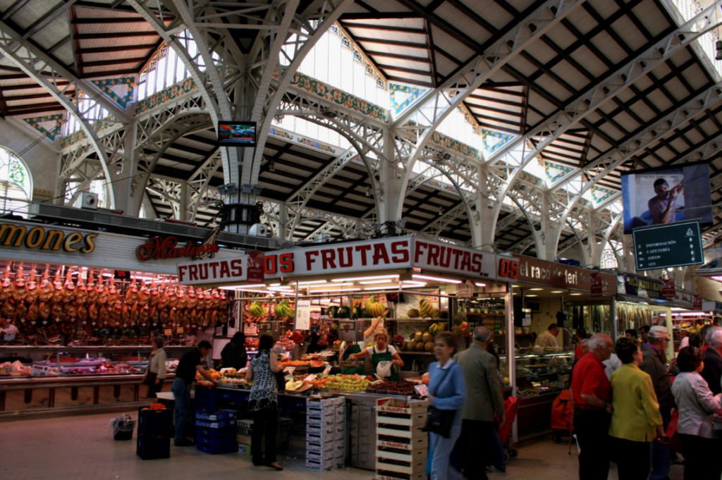 Indoor view of a bustling market with various fruit and meat stalls, shoppers browsing, and an ornate iron and glass ceiling above.