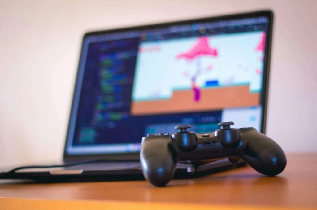 Video game controller in focus on a desk with a blurred laptop screen displaying colorful graphics in the background.