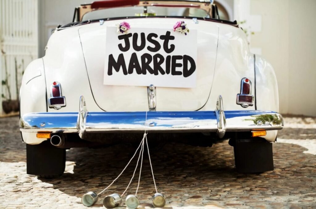 White vintage car with "Just Married" sign and tin cans tied to the rear bumper.