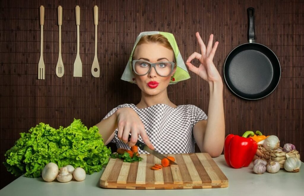 Woman in vintage kitchen attire making an OK gesture with one hand while chopping carrots with the other, surrounded by various vegetables and cooking utensils, with a frying pan hanging on the wall behind her.