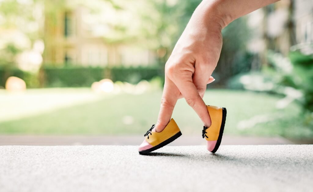 A person's fingers are posed to look like legs walking, with miniature shoes on the fingertips, against a blurred outdoor background.