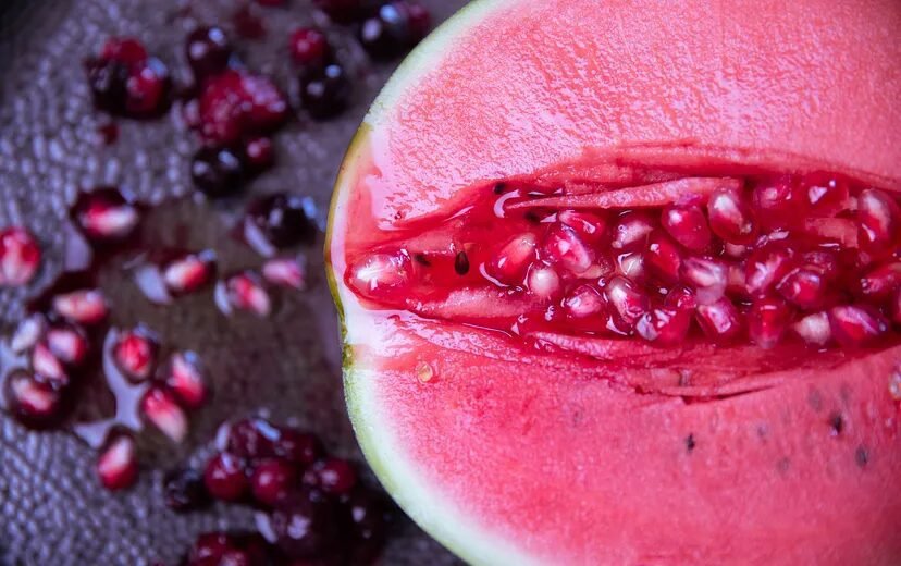 Half of a watermelon with a section carved out, revealing both the red flesh and the seed-filled center, with loose seeds scattered on the surface beneath.
