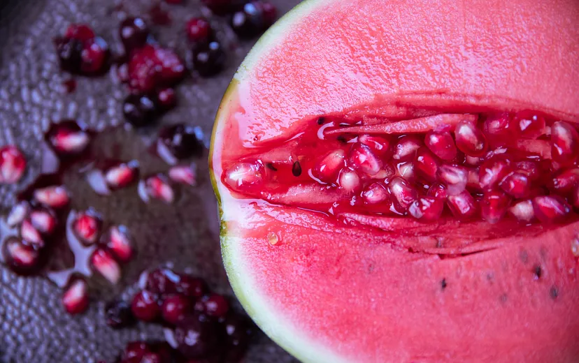 Half of a watermelon with a section carved out, revealing both the red flesh and the seed-filled center, with loose seeds scattered on the surface beneath.