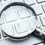 Close-up of white keyboard with magnifying glass on G key, symbolizing detailed digital examination.