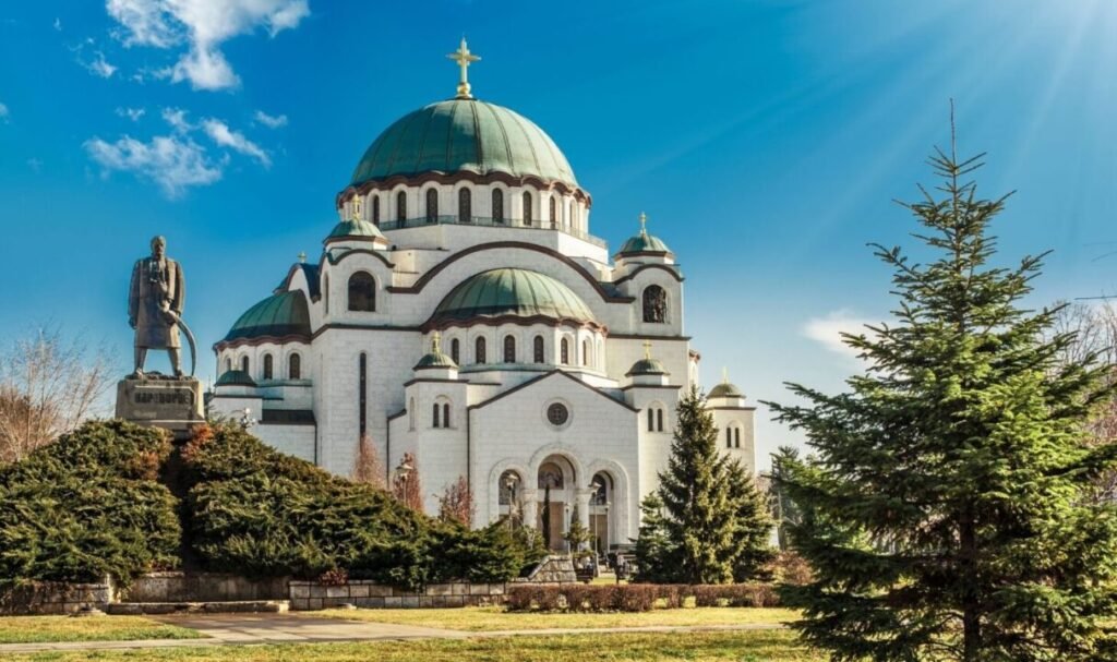 Alt text: A large white Orthodox church with green domes under a clear blue sky, sunbeams shining through on the right, and a statue in front surrounded by greenery.