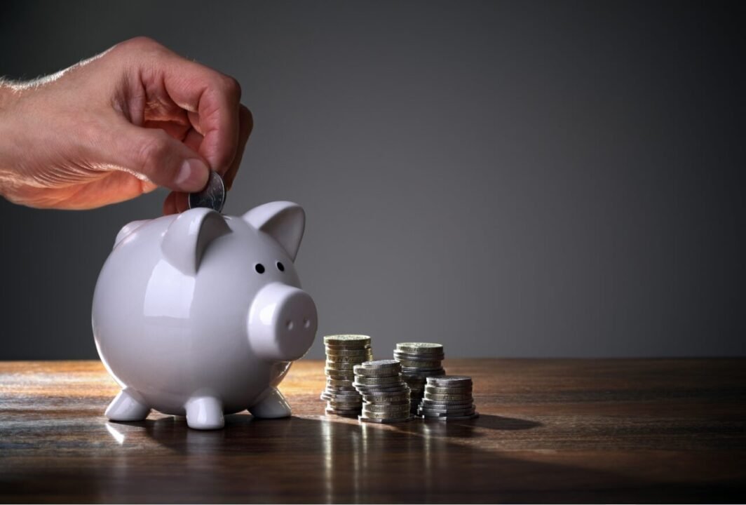 Saving money: hand adding coin to white piggy bank with stacks of coins on wood.