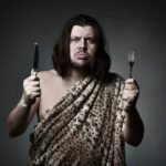 Wild hunter in animal print with knife and fork in comical mealtime standoff.