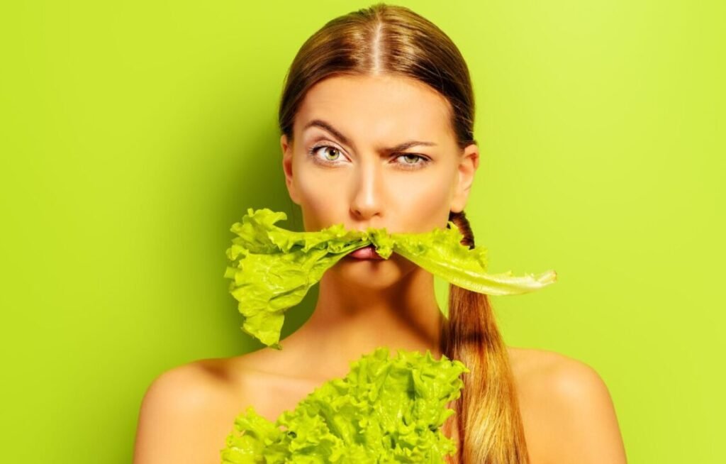 A woman holding a leaf of lettuce in her mouth against a lime green background.