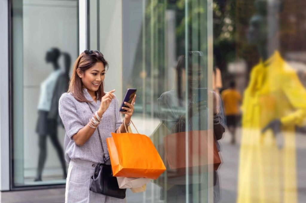 Woman happily using her smartphone while holding shopping bags on a city street.