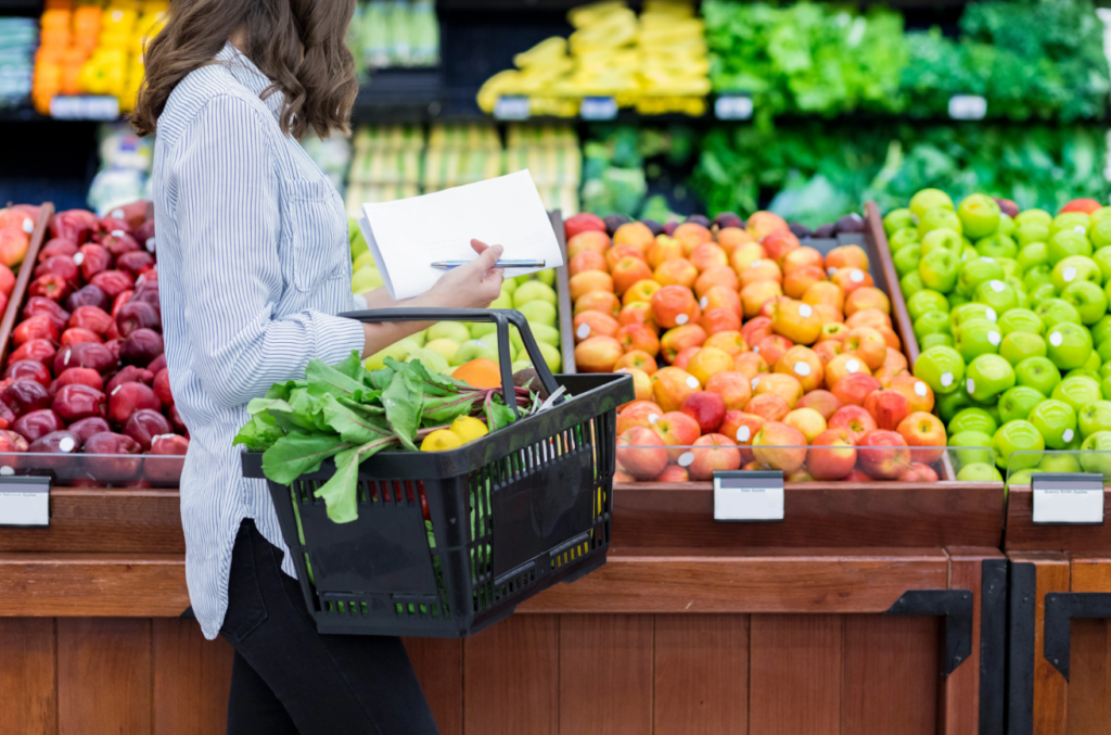 A woman in a striped shirt holding a shopping basket filled with vegetables and a notepad, standing next to a display of colorful fruits in a grocery store.