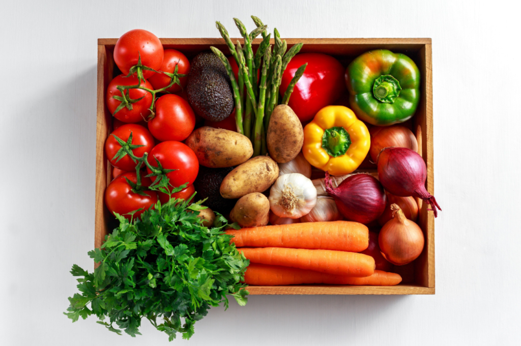 A variety of fresh vegetables including tomatoes, avocados, asparagus, bell peppers, potatoes, garlic, onions, and carrots neatly arranged in a wooden box on a white surface.