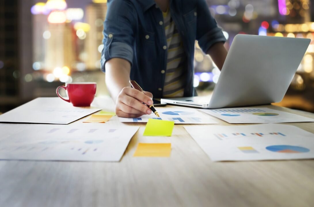 Person working at a table with sticky notes, charts, a red cup, and a laptop, with blurry city lights in the background.