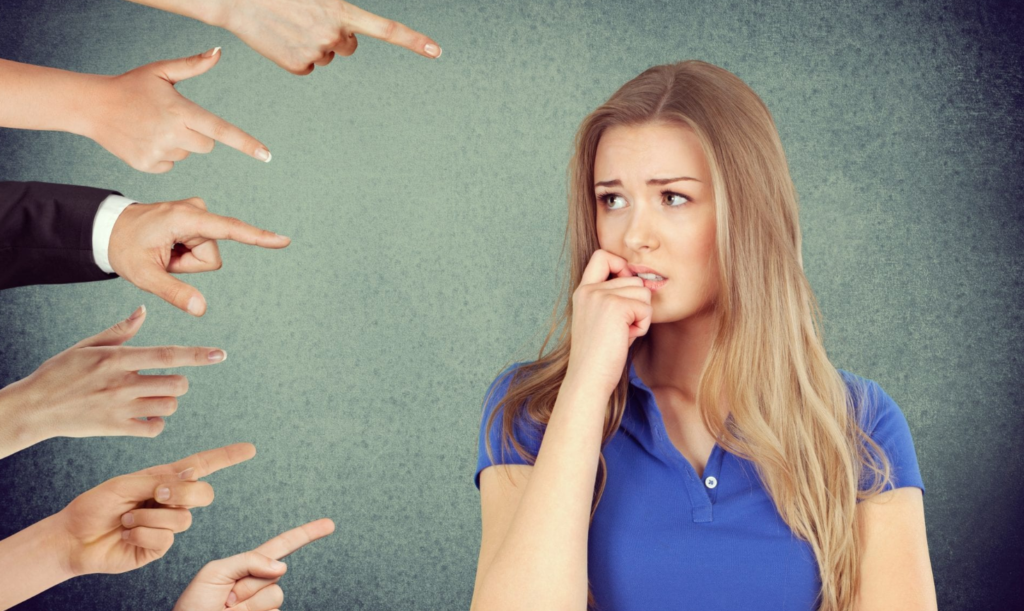 A worried-looking young woman with her hand on her chin, surrounded by multiple hands pointing at her, representing accusation or blame, against a textured green background.