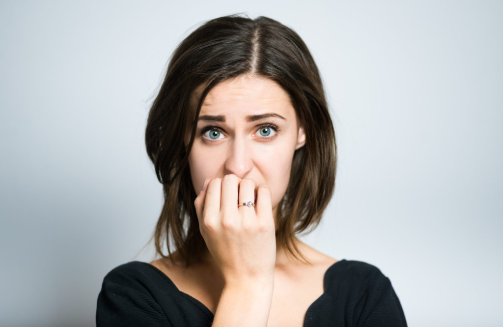 A worried-looking young woman with her hand covering her mouth against a plain background.