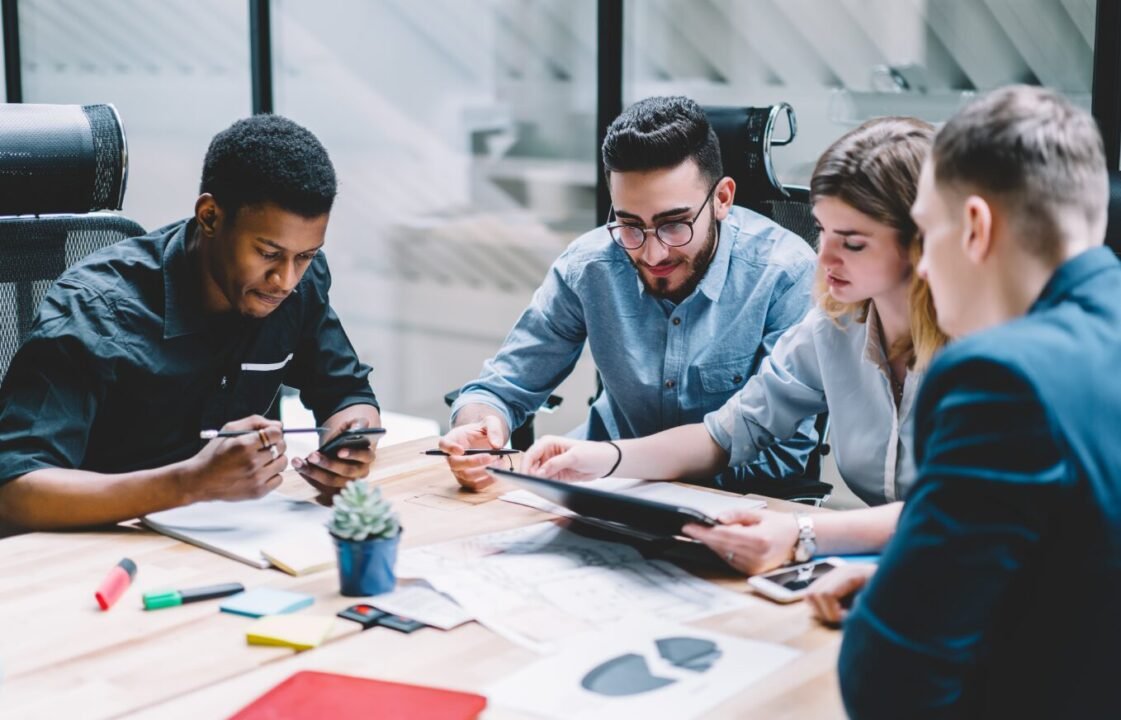 Group of diverse young professionals collaborating at a table with papers and digital devices in a modern office setting.