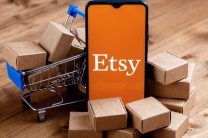 Smartphone displaying the Etsy logo surrounded by small cardboard boxes and a miniature shopping cart.