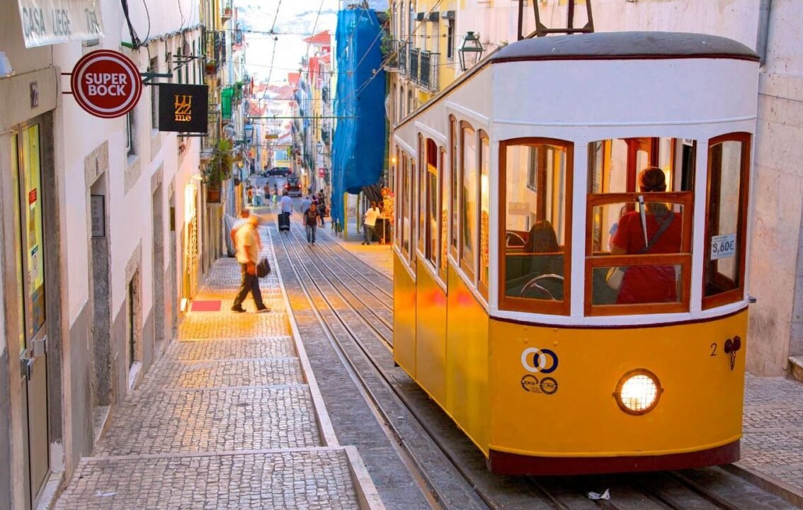 A yellow tram ascends a steep, cobbled street in a European city, with pedestrians walking alongside.