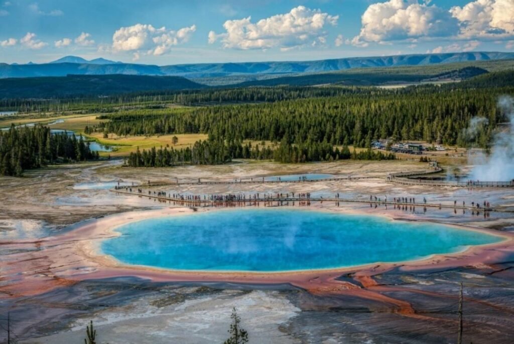 Aerial view of the Grand Prismatic Spring in Yellowstone National Park, surrounded by lush forests and distant mountains, with a boardwalk filled with tourists encircling the vibrant, colorful hot spring.