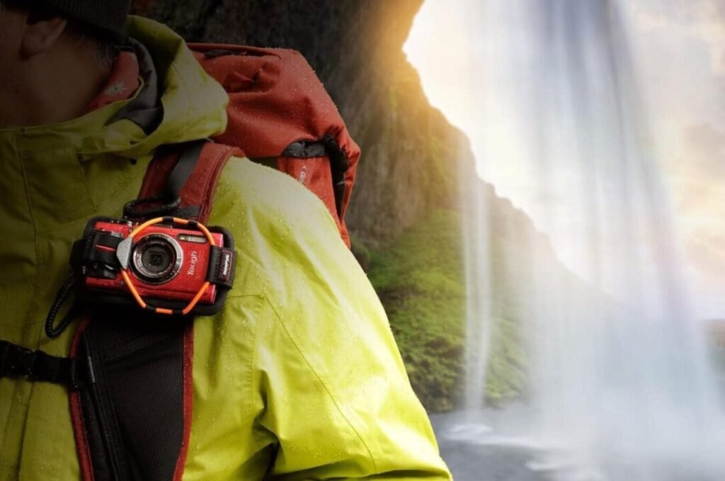 Close-up of a person wearing a bright yellow jacket with a red camera attached, standing near a waterfall with sunlight filtering through mist.