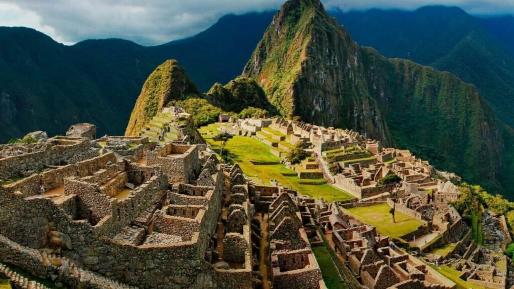 Aerial view of Machu Picchu, an ancient Inca city with well-preserved ruins and lush green terraces, nestled between two mountain peaks under a partly cloudy sky.