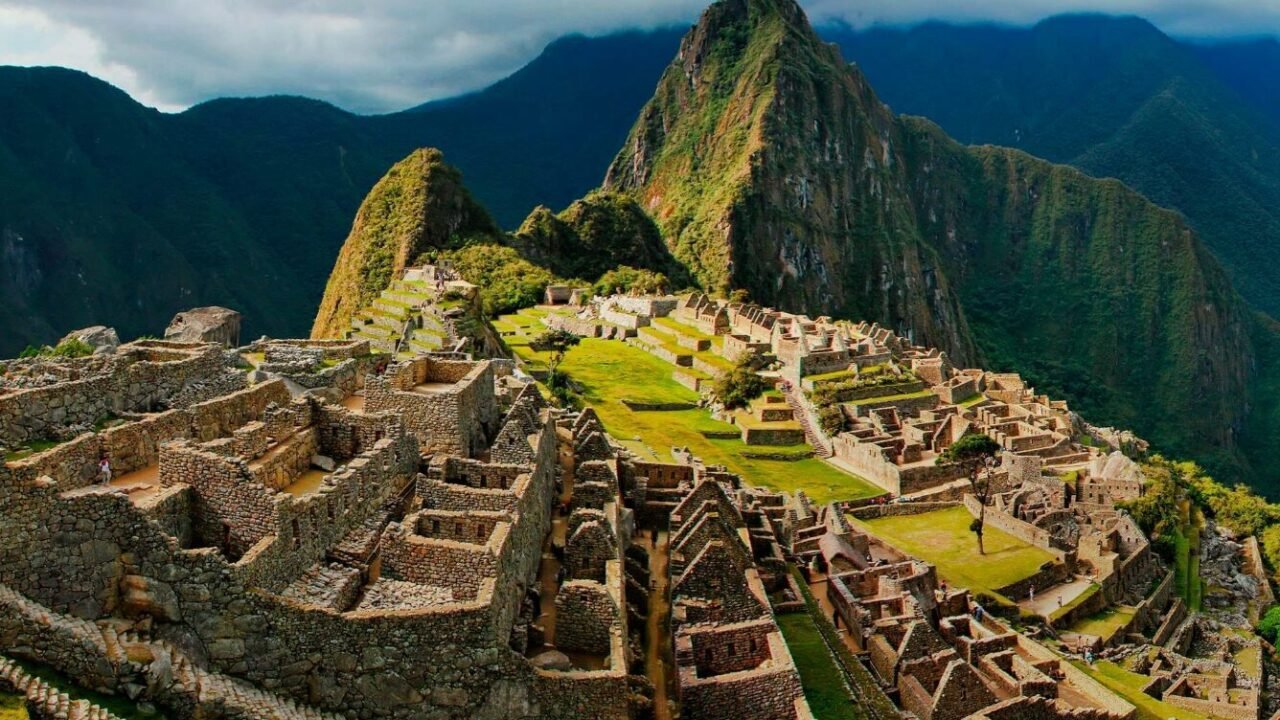 Aerial view of Machu Picchu, an ancient Inca city with well-preserved ruins and lush green terraces, nestled between two mountain peaks under a partly cloudy sky.