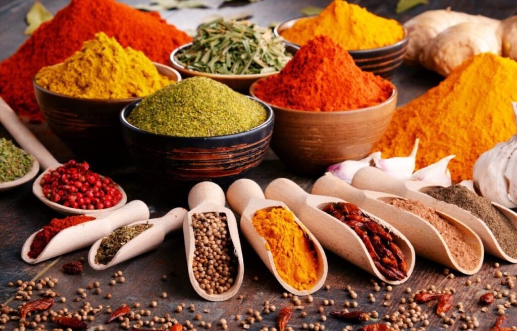 Assorted spices in bowls and wooden scoops on a table, including red, yellow, and green powders, whole red chilies, and garlic cloves.