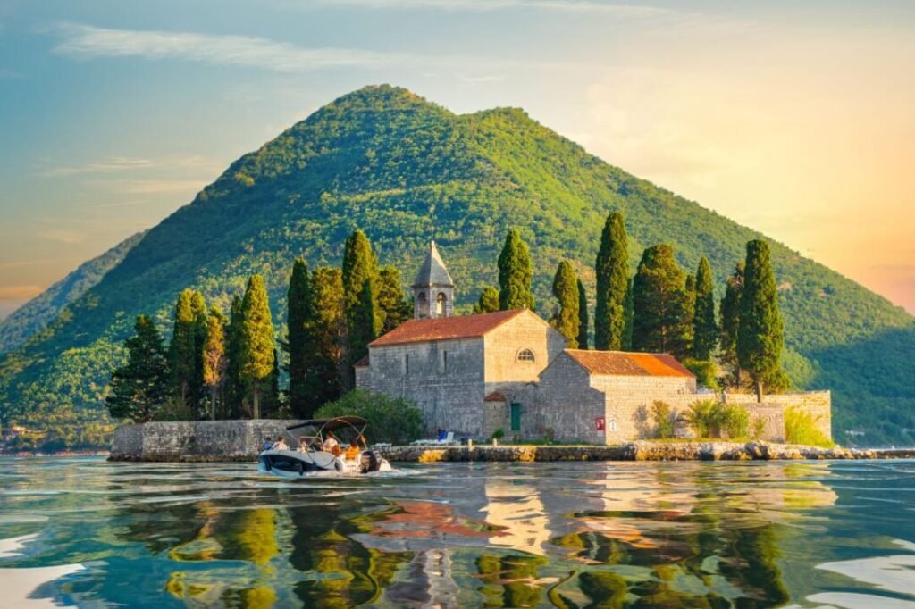 Stone church on a small island  in Montenegro with tall cypress trees in front of a lush green mountain, with a boat in the foreground on a calm body of water.