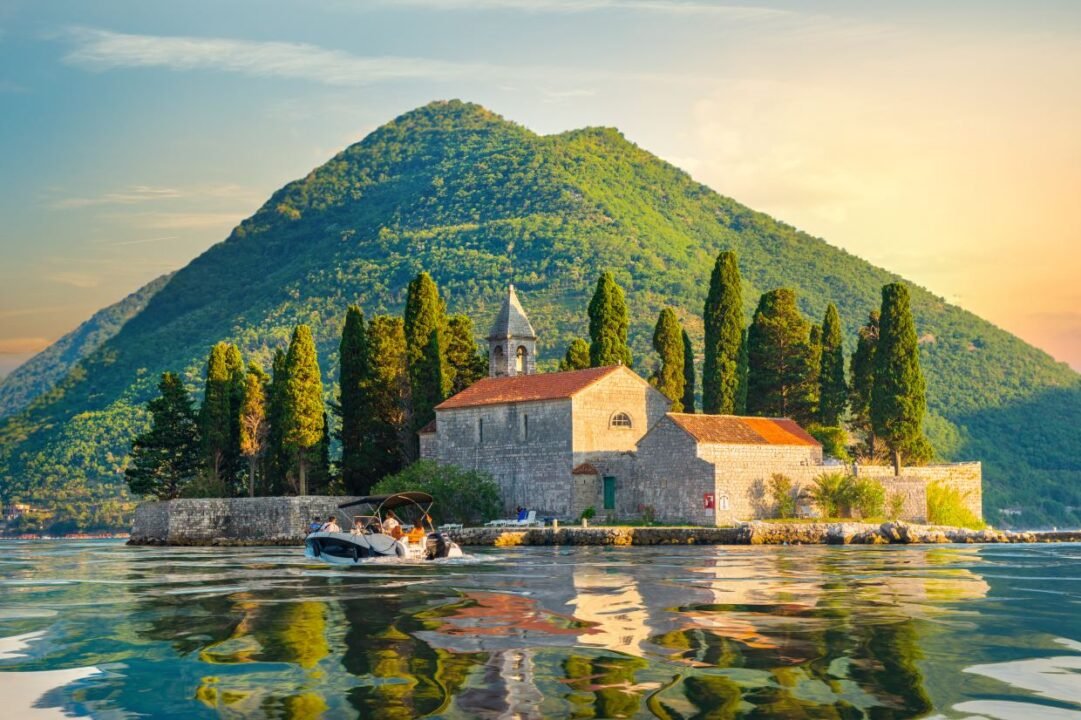 A stone building with a red-tiled roof, surrounded by tall trees, on a small island n Montenegro with a mountain in the background and a boat on the water.