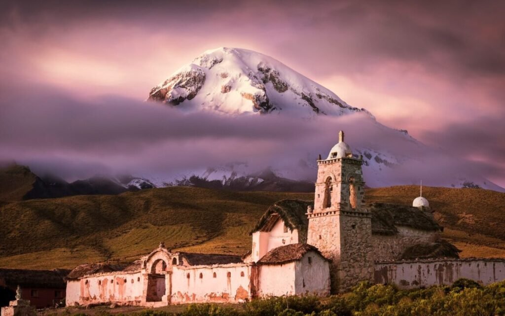 Old church with bell tower in the foreground set against a backdrop of a snow-capped mountain and vibrant pink clouds at sunset.