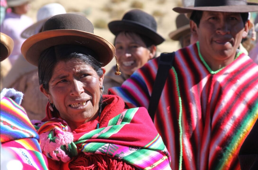 Indigenous Bolivian woman in traditional attire including a bright red poncho and a brown bowler hat, with two smiling people in the background wearing similar cultural outfits.