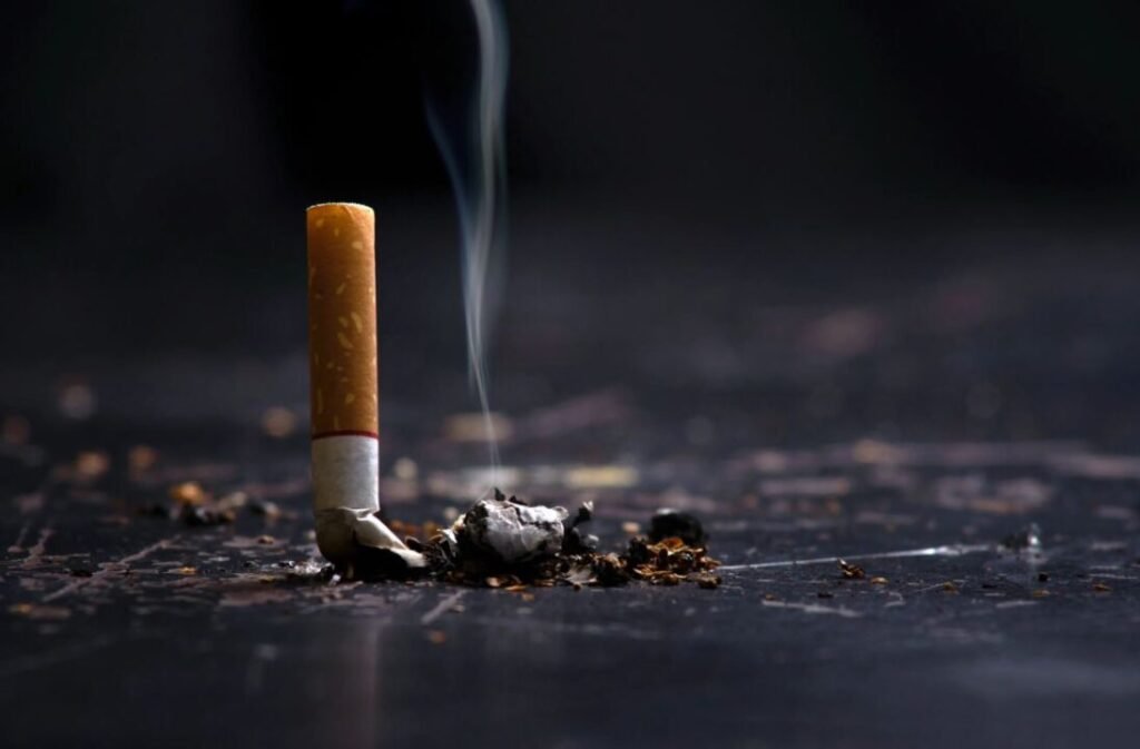 A cigarette butt with smoke rising from it, lying on a dark surface scattered with ashes.